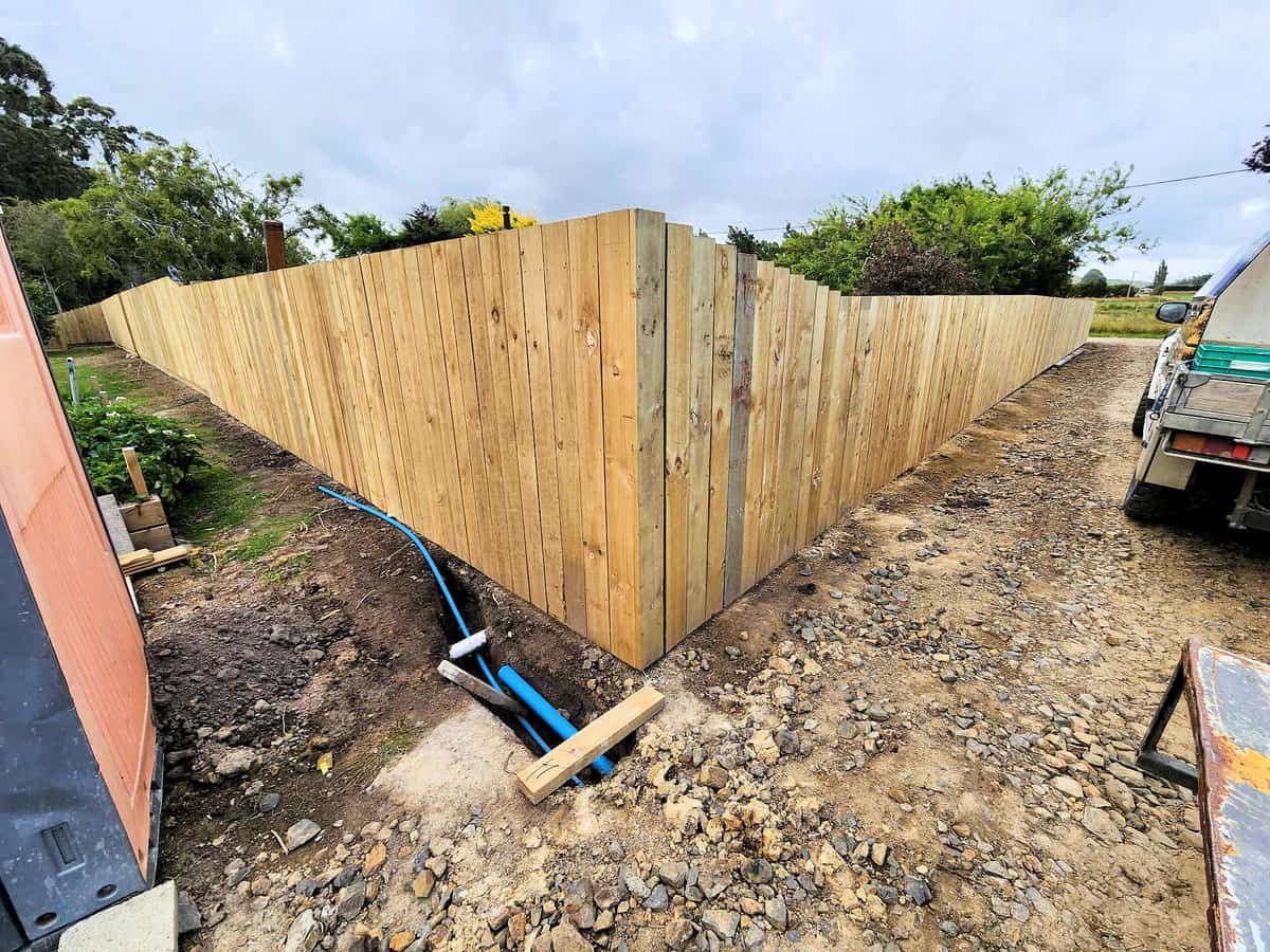 Tall timber paling boundary fence running down both sides of a new driveway, with a service trench and blue water main open along the fence line