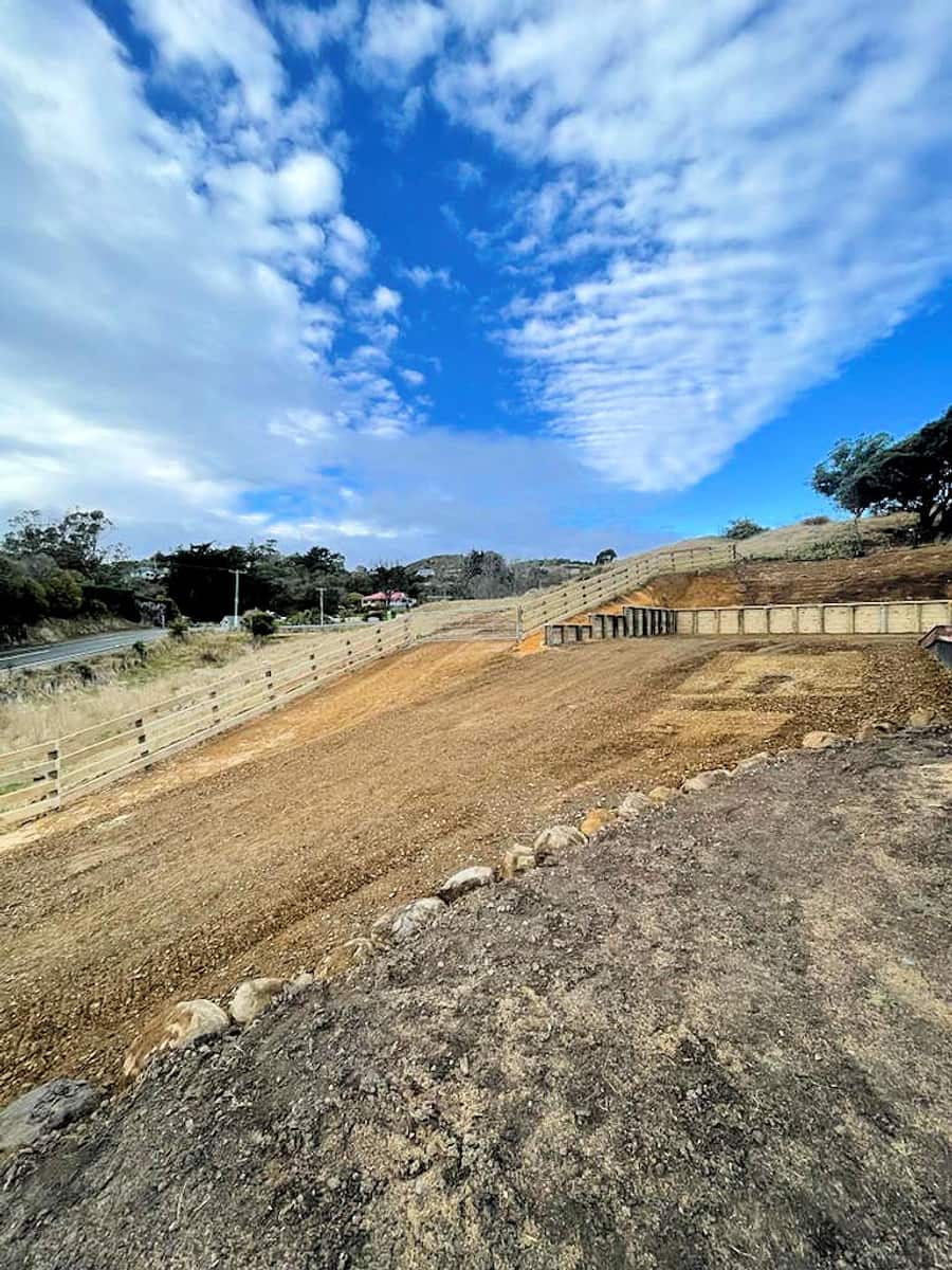 Cut-and-fill building platform on a coastal hillside with a timber post and rail barrier fence running the length of the batter above a state highway