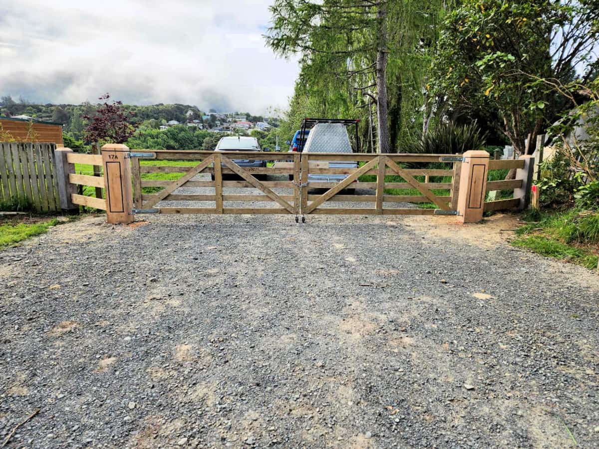 Wide timber farm gateway with capped strainer posts and a gravel entrance, framed by trees