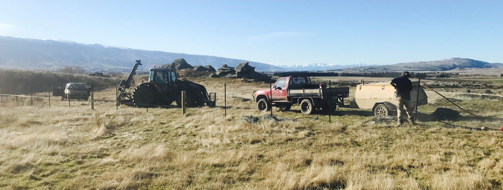 Tractors, a ute and a wire trailer working a long fence line across dry tussock country with hills behind