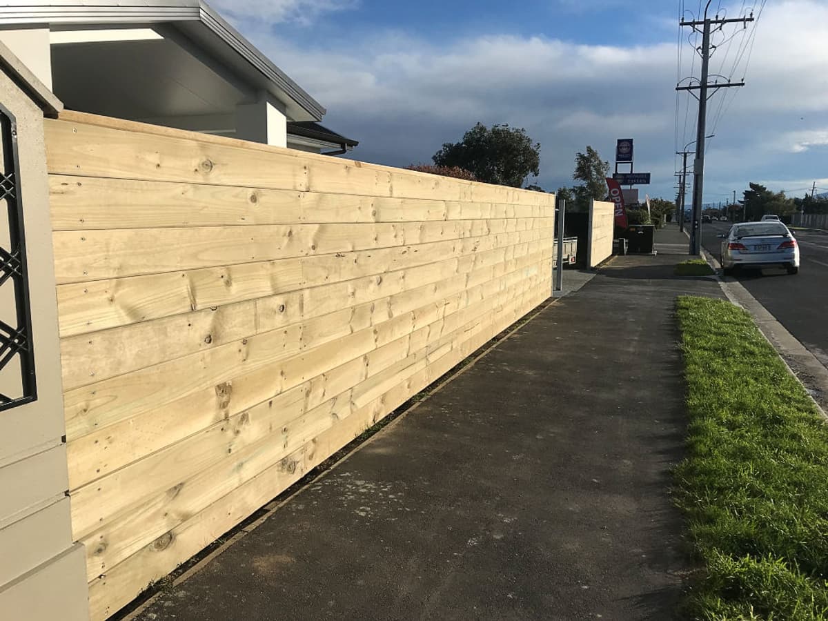 Horizontal timber slat boundary wall running down a commercial street frontage beside a concrete footpath, with parked cars and power poles beyond