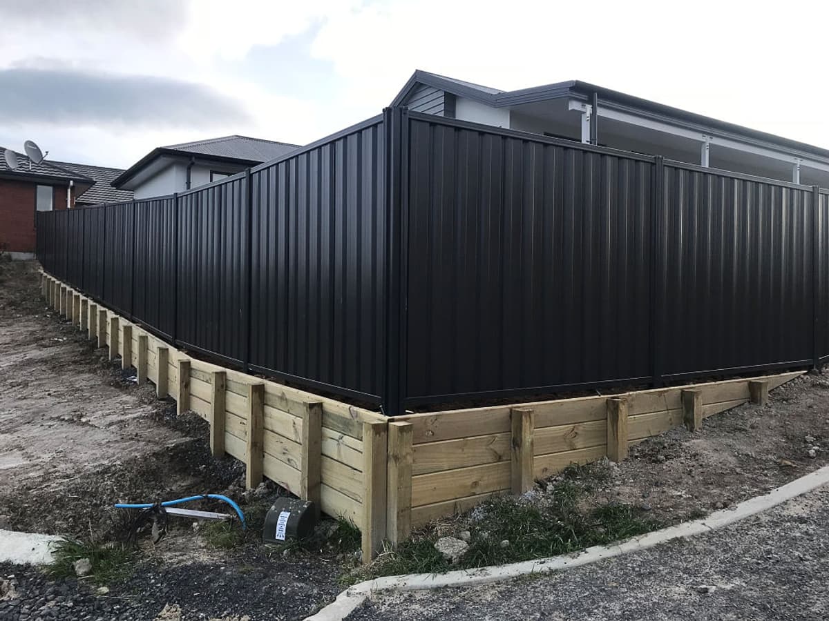 Dark colour-steel boundary fence built on a timber retaining wall around the corner of a new-build house, with kerb, channel and services still exposed