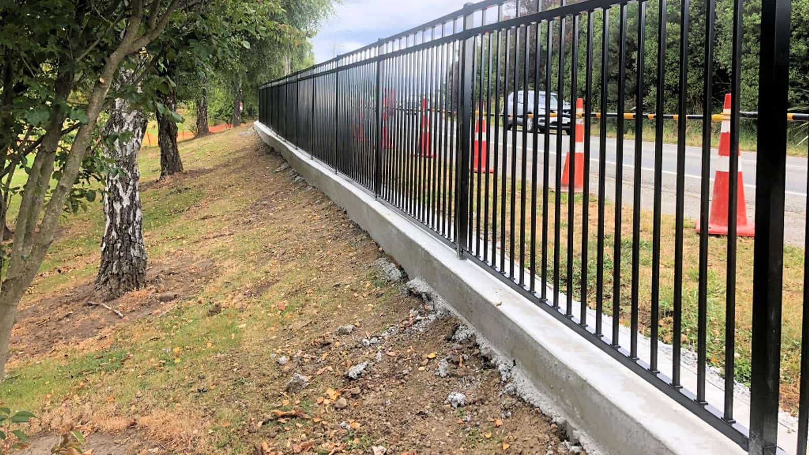 Black steel palisade security fence on a poured concrete nib running beside a public road, with road cones and traffic management set out along the berm